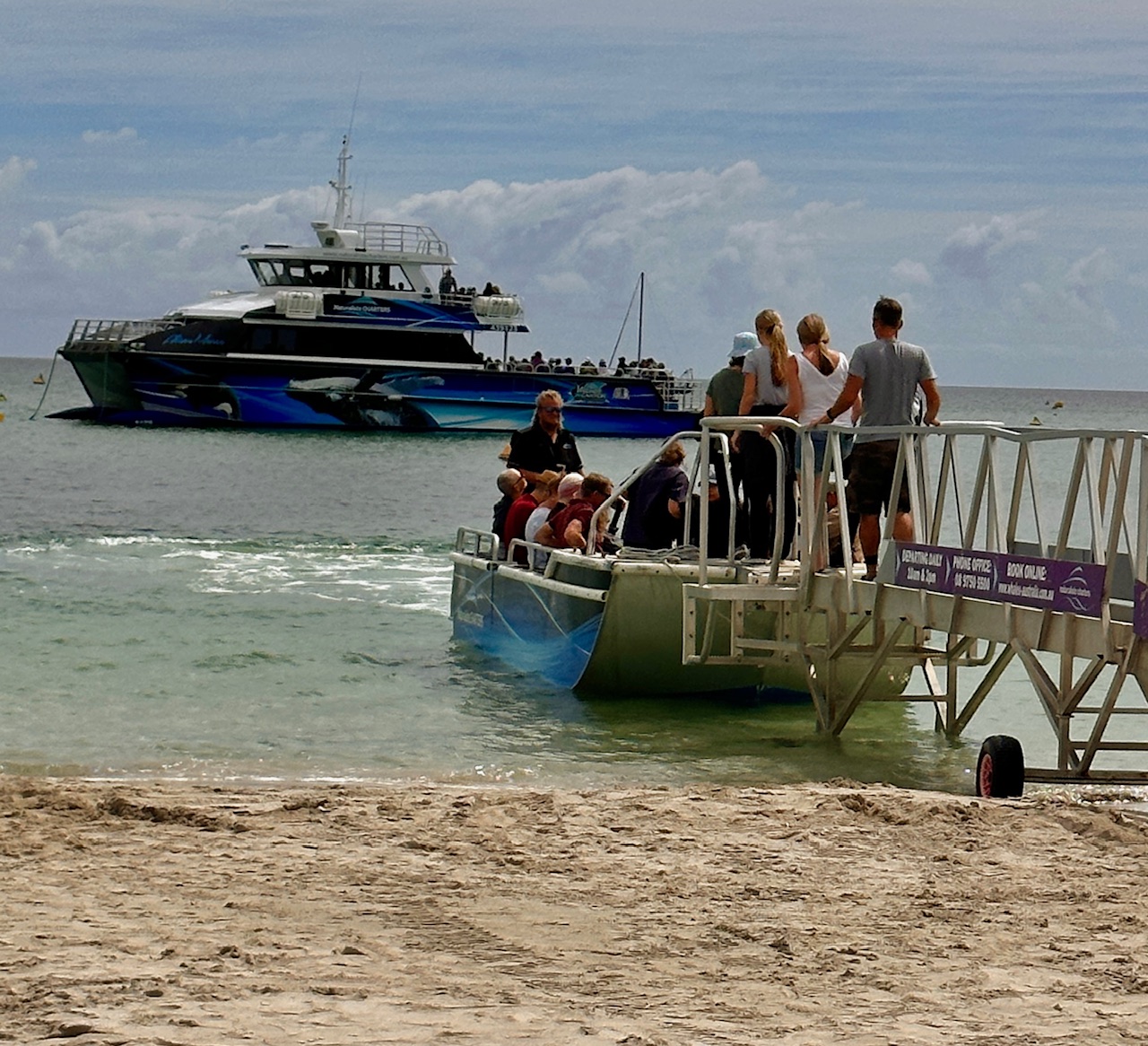 An image of people boarding a beachside ramp arching into the ocean, a small tender is collecting them to take them across to the Alison Maree whale watching boat moored further out to sea.